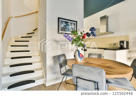 A stylish modern interior featuring a gracefully curved staircase, a round dining table, contemporary chairs, and a vibrant floral centerpiece enhancing the space. 125502448