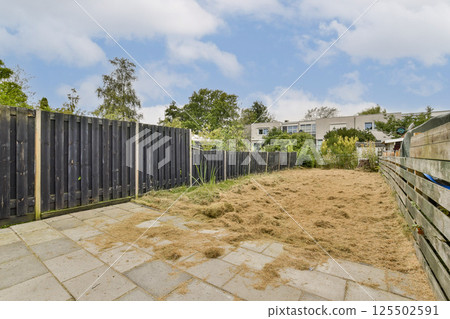 A serene, untended garden is surrounded by wooden fencing, creating a peaceful outdoor space. The area has patches of overgrown grass and sparse vegetation. A serene, untended garden is surrounded by wooden fencing, creating a peaceful outdoor space. The area has patches of overgrown grass and sparse vegetation. 125502591