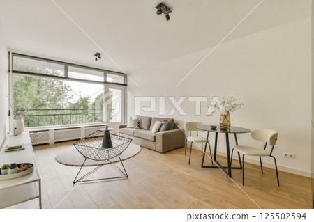 A stylish minimalist living room featuring a neutral color palette, modern furniture, and ample natural light from large windows enhancing the inviting atmosphere. 125502594