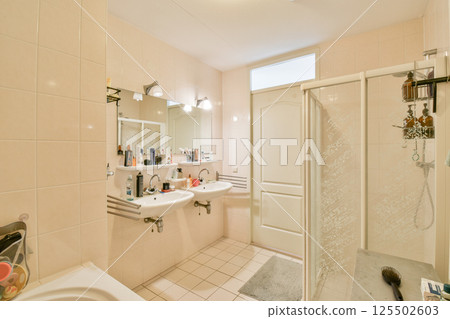 A spacious and contemporary bathroom featuring two sinks, mirrors, and a glass shower. The light-colored tiles and natural light create an inviting atmosphere. 125502603