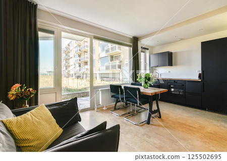 A stylish and modern apartment interior featuring large windows, a dining area, and sleek kitchen design emphasizes comfort and elegance. 125502695