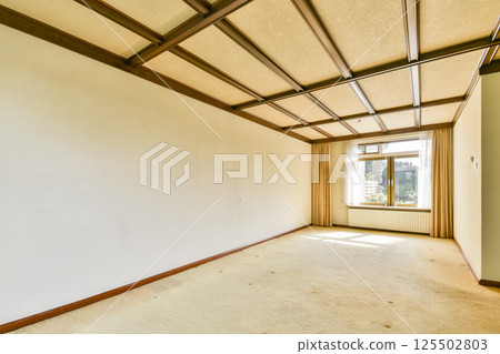 An empty room featuring a minimalist design with a large window and warm lighting. The ceiling showcases sleek lines and modern architecture. 125502803