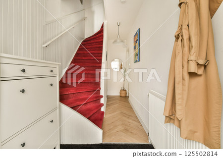 A stylish contemporary staircase adorned with a red carpet, leading up in a well-lit hallway featuring minimalistic decor and elegant furniture. 125502814