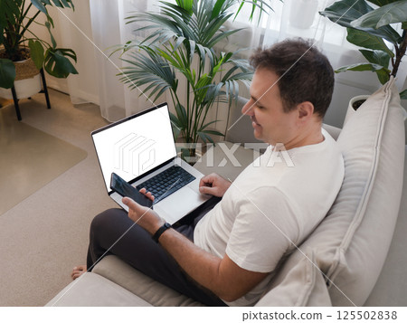 Smiling man sitting with laptop on knees, looking at smartphone. Laptop screen is visible, ideal for mockup use. Concept of positive digital interaction, remote work, and modern technology lifestyle. 125502838