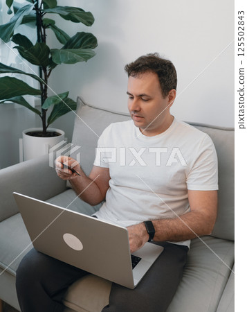 Side view of a man holding a bank card and verifying details in a modern living room with a fiddle leaf fig plant. Concept of secure online payment, digital banking, e-commerce and online shopping 125502843