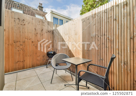 A serene outdoor patio features wooden fencing, a small table with chairs, and a barbecue grill, perfect for relaxation and gatherings. A serene outdoor patio features wooden fencing, a small table with chairs, and a barbecue grill, perfect for relaxation and gatherings. 125502871