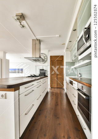 A sleek, modern kitchen featuring an open layout with wooden floors, white cabinetry, and stainless steel appliances, ideal for contemporary living spaces. 125502874