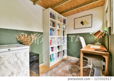 A stylish and organized workspace featuring a bookshelf, desk lamp, and minimalistic decor. The space is bright and inviting, ideal for productivity. 125502917