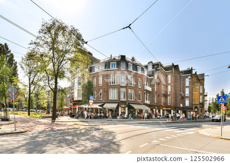 A charming street scene featuring historic buildings, vibrant greenery, and pedestrians enjoying a sunny day. The architecture exhibits a mix of styles. 125502966