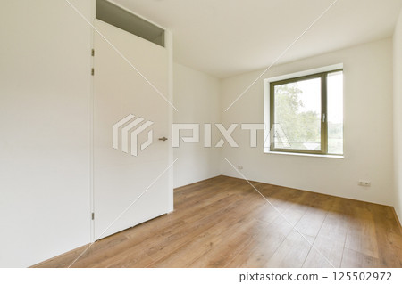 A spacious minimalist interior featuring white walls and a large window, allowing ample natural light to illuminate the wooden floor. A spacious minimalist interior featuring white walls and a large window, allowing ample natural light to illuminate the wooden floor. 125502972