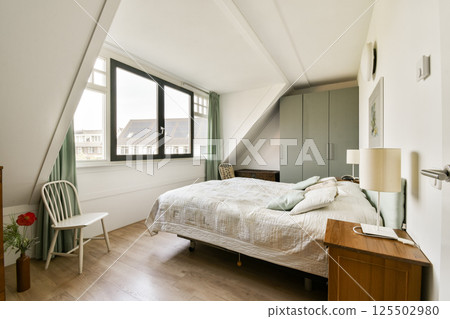 A beautifully designed modern bedroom featuring a cozy bed, natural light streaming through large windows, and minimalist decor for a serene atmosphere. 125502980