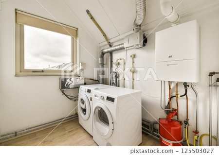 Bright and functional laundry room featuring modern washing machines, water pipes, and ample natural light from a window. 125503027