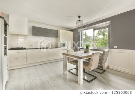 A stylish modern kitchen featuring sleek cabinetry, a dining table, and ample natural light from a large window, creating an inviting atmosphere. 125503068