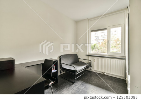 A minimalist room featuring a desk and chair, accompanied by a comfortable seat beside a large window providing natural light and an outside view. 125503083