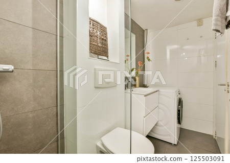 A stylish modern bathroom featuring a glass shower, toilet, washing machine, and elegant floral arrangements that enhance the decor. A stylish modern bathroom featuring a glass shower, toilet, washing machine, and elegant floral arrangements that enhance the decor. 125503091