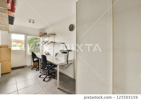A stylish modern office space designed to maximize natural light, featuring a clean workspace with desks, shelves, and a large window overlooking greenery. 125503159
