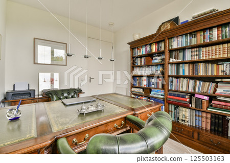 A sophisticated home office featuring a polished wooden desk, comfortable green leather chairs, and a large bookshelf filled with books, creating a cozy work atmosphere. 125503163