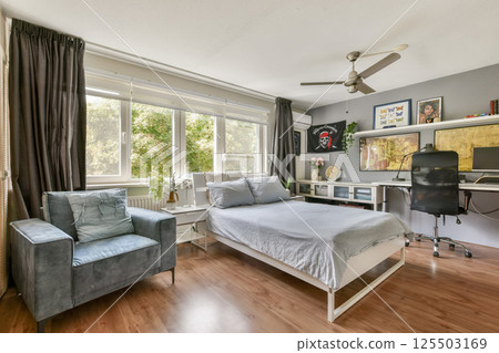 A stylish bedroom featuring a soft gray bed, a cozy armchair, and a practical workspace by the window, surrounded by natural light. A stylish bedroom featuring a soft gray bed, a cozy armchair, and a practical workspace by the window, surrounded by natural light. 125503169