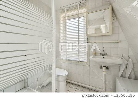 A sleek and modern minimalist bathroom featuring white tiled walls, a contemporary sink, and a stylish shower with frosted glass. Natural light pours in from a window. 125503190