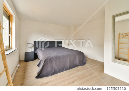 A serene minimalistic bedroom featuring a bed with a gray coverlet, a wooden ladder, a mirror, and natural light illuminating the space. 125503210