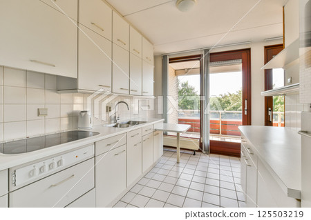 A bright and spacious kitchen featuring sleek white cabinets, a tiled floor, and large windows offering a view of the outdoors. 125503219