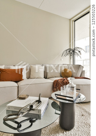 A stylish modern living room featuring a cozy sofa, decorative cushions, a sleek coffee table, and large window allowing natural light, ideal for contemporary living. A stylish modern living room featuring a cozy sofa, decorative cushions, a sleek coffee table, and large window allowing natural light, ideal for contemporary living. 125503226