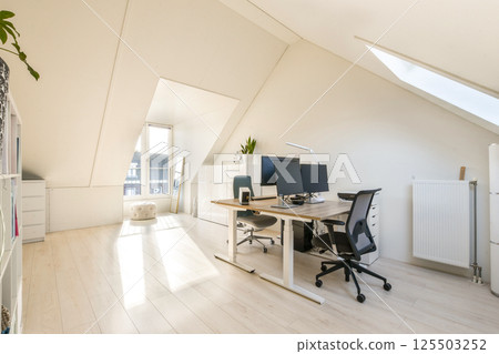 Bright and airy modern office space featuring sleek furniture, dual computer setups, and large windows allowing ample sunlight. A stylish, minimalistic design adds to productivity. 125503252