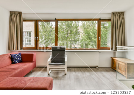 A contemporary living room featuring a red sofa, wooden furniture, and large windows overlooking greenery. The space is well-lit and inviting, blending comfort with style. 125503527