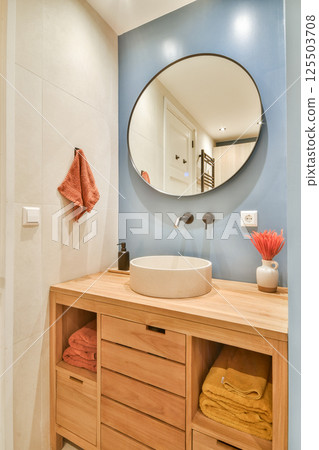 An aesthetically pleasing modern bathroom featuring a circular mirror, stylish sink, and wooden cabinetry, accented by vibrant towels. An aesthetically pleasing modern bathroom featuring a circular mirror, stylish sink, and wooden cabinetry, accented by vibrant towels. 125503708
