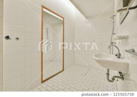 A spacious, minimalist bathroom featuring white tiled walls and floor, a large mirror, and sleek plumbing fixtures. The uncluttered design offers a clean, serene environment. 125503739