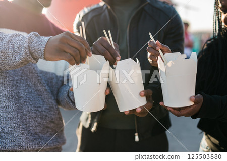 Three african american friends enjoying Asian street food outdoors on a sunny day, using chopsticks and sharing casual moments near a bright orange food truck. Warm backlight creates a cozy, friendly 125503880
