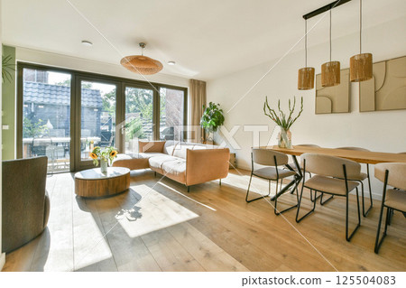 A bright and stylish living area featuring a comfortable sofa, wooden dining table, and large windows that invite natural light. Perfect for modern living aesthetics. 125504083