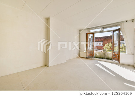 A sunlit, empty room featuring white walls and large windows that invite natural light, providing an airy and spacious atmosphere. 125504109