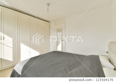 A bright and spacious modern bedroom featuring minimalistic design with white cabinetry and a comfortable bed dressed in gray linens. Ideal for contemporary living. 125504170
