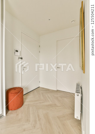 A contemporary entrance hallway featuring a stylish round ottoman, sleek doors, and minimalistic decor. The warm tones and clean lines create an inviting atmosphere. 125504211