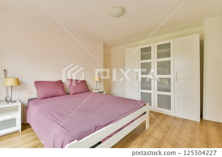 A contemporary bedroom featuring a cozy bed with purple bedding, minimalist decor, and a soothing color palette. Ideal for relaxation and comfort. 125504227