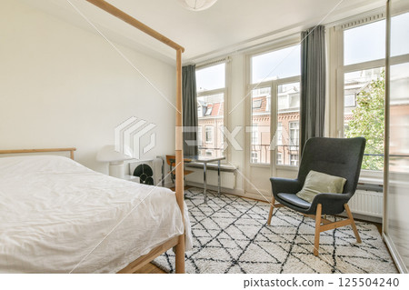 A bright, airy bedroom featuring a wooden bed, modern armchair, and natural light streaming through large windows, creating a serene atmosphere. 125504240