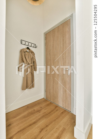 An elegant entryway featuring a minimalist design, wooden flooring, and a coat hanging on a wall rack. The aesthetic blends functionality with modern style. 125504295