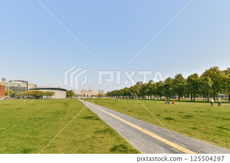 An expansive urban park featuring well-maintained pathways, lush green grass, and scattered visitors enjoying the serene environment and pleasant weather. An expansive urban park featuring well-maintained pathways, lush green grass, and scattered visitors enjoying the serene environment and pleasant weather. 125504297