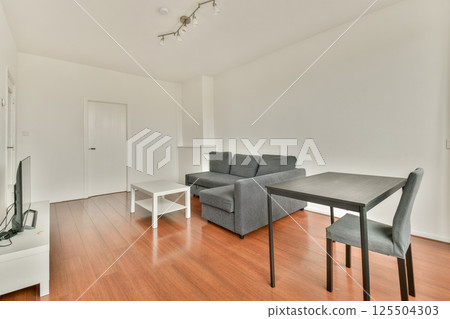 A spacious and modern living room features a grey couch, minimalist furniture, and wooden flooring, creating a stylish and inviting atmosphere for relaxation. 125504303