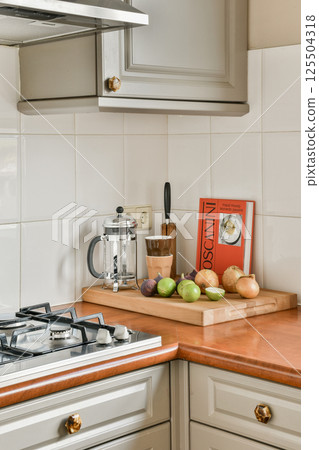 A bright, modern kitchen showcases a wooden countertop with fresh fruits and vegetables, a kettle, and a cookbook, creating a warm cooking atmosphere. 125504318