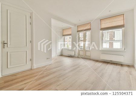 A spacious, modern living room with light-colored walls, wooden flooring, and large windows dressed with minimalistic blinds. 125504371