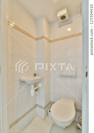 A clean and minimalist bathroom showcasing white tiled walls and floor, a small sink, and a toilet. Simple design emphasizing functionality and space. 125504410