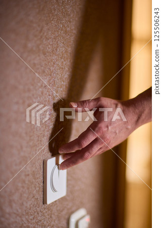 A hand reaches for the light switch in a cozy room, adding warmth and brightness A hand reaches for the light switch in a cozy room, adding warmth and brightness 125506243
