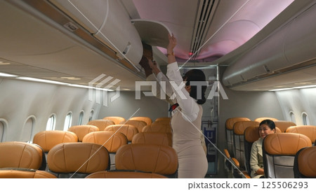 Friendly flight attendant. Air hostage helping passenger businessman put luggage on top shelf or cabin compartment on airplane, woman help black man lift suitcases putting overhead locker on airplane Friendly flight attendant. Air hostage helping passenger businessman put luggage on top shelf or cabin compartment on airplane, woman help black man lift suitcases putting overhead locker on airplane 125506293