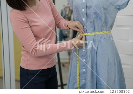 A woman measuring a dress on a mannequin for tailoring or design, showcasing fashion and craftsmanship. 125508502