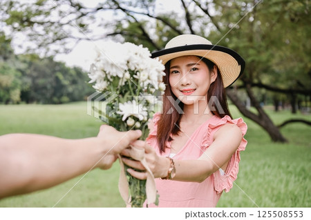 A young woman in a hat offers a bouquet of flowers, smiling warmly in a lush green park. 125508553