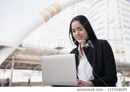 A professional woman enjoys working on her laptop outside in a modern urban setting, symbolizing business success. A professional woman enjoys working on her laptop outside in a modern urban setting, symbolizing business success. 125508609