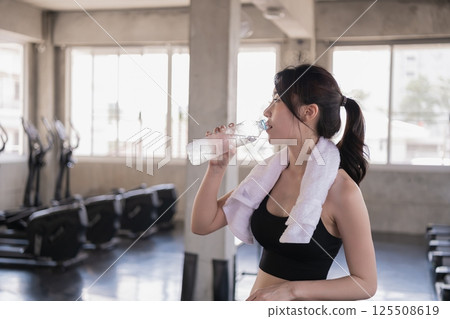 A woman in a gym drinks water after an intense workout, showcasing the importance of hydration and fitness. A woman in a gym drinks water after an intense workout, showcasing the importance of hydration and fitness. 125508619