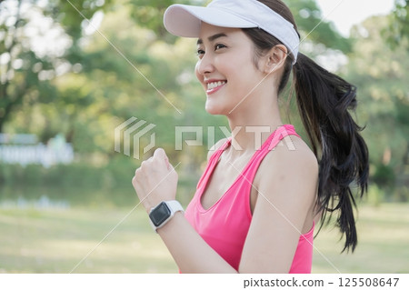 A joyful woman jogging in a park, wearing sporty attire and a smartwatch, embracing a healthy lifestyle. 125508647